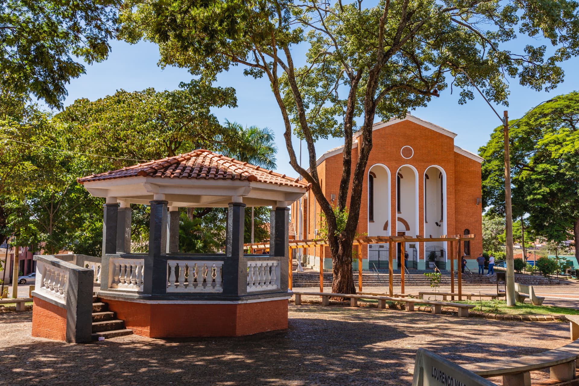 Coreto da Praça da Igreja Matriz de Serrana, com a igreja de tijolos aparentes ao fundo, árvores e bancos sob céu azul